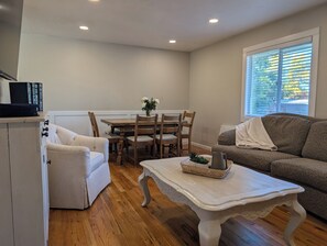 A cozy living room with a wooden dining table, two chairs, a white sofa, and a white armchair. A coffee table with a basket and a pitcher is in the center.