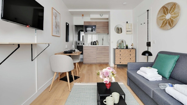 A modern living room with a flat-screen TV, a grey sofa, a coffee table with a vase of flowers, and a kitchen in the background. - Peaceful Studio -2P- Moulin Rouge (Paris)