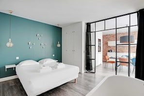 A modern hotel room with a large bed, a view of the kitchen through a glass door, and a balcony with chairs.
