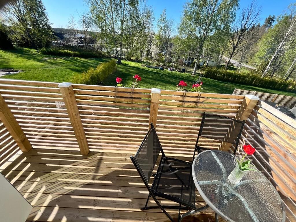A wooden deck with a glass table and chairs, surrounded by a wooden fence, overlooking a green landscape with trees and a clear sky.