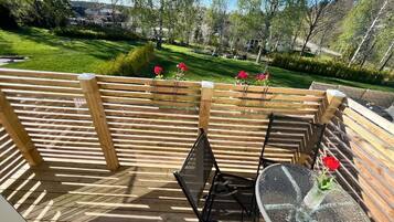 A wooden deck with a glass table and chairs, surrounded by a wooden fence, overlooking a green landscape with trees and a clear sky.