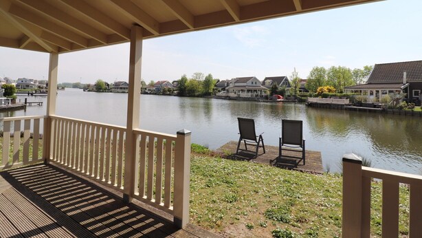 Eine überdachte Terrasse mit Blick auf einen ruhigen Fluss, zwei Stühle und ein Holzsteg.