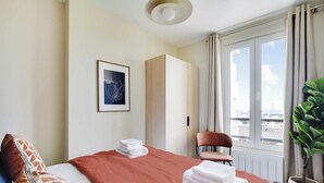 Habitación de hotel con cama, una silla, ventana con cortinas, planta en maceta y un cuadro en la pared.