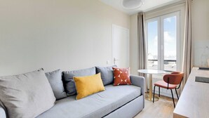Una sala de estar moderna con un sofá gris, un cojín amarillo y un cojín rojo estampado. Se observa una mesita redonda con una silla, una puerta blanca y una ventana con cortinas.