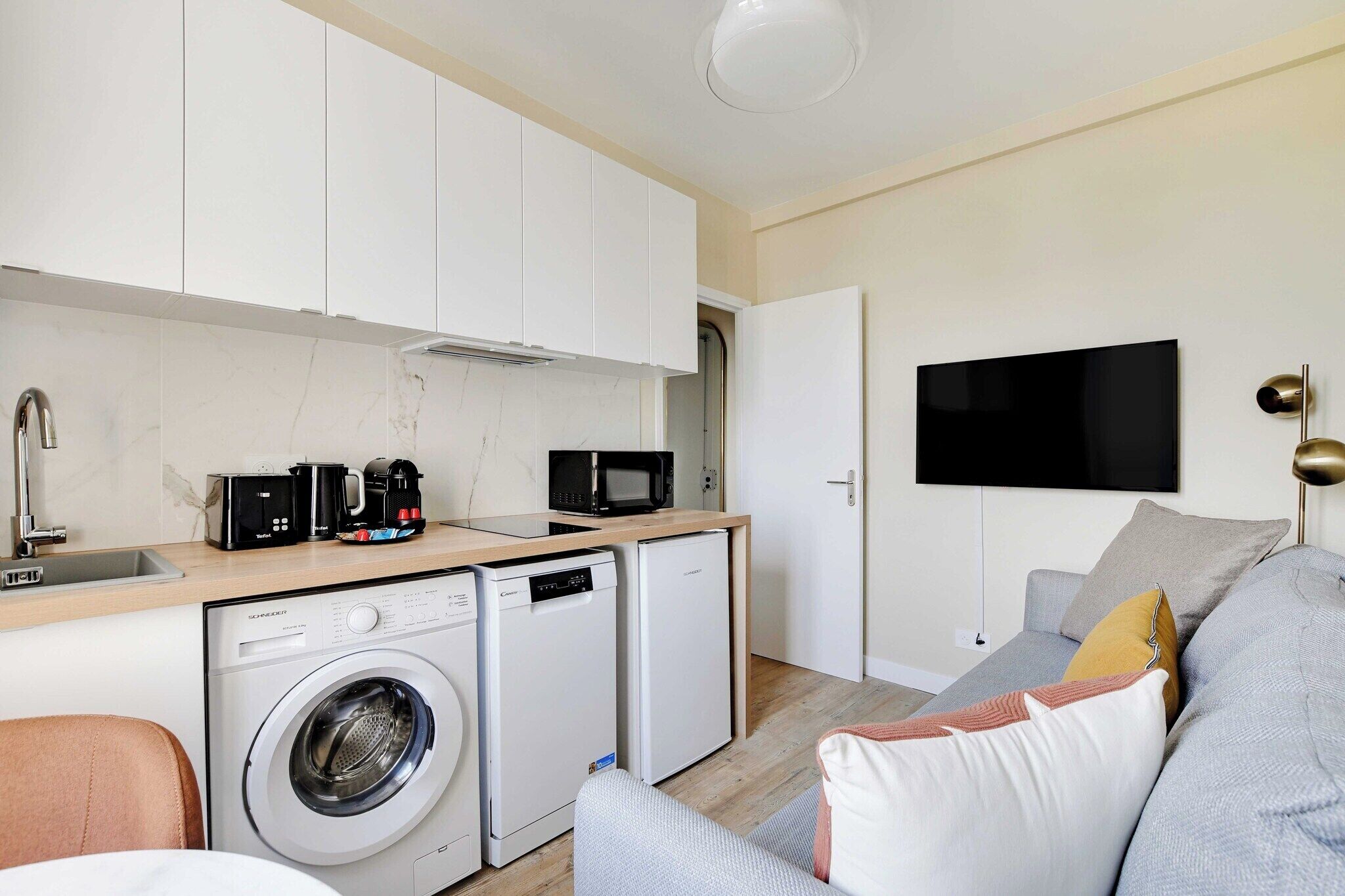 A compact living space with a washing machine, a compact refrigerator, a microwave, a coffee maker, and a flat-screen TV mounted on the wall.