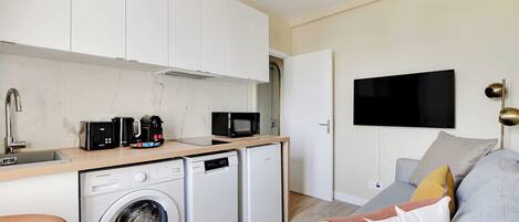 A compact living space with a washing machine, a compact refrigerator, a microwave, a coffee maker, and a flat-screen TV mounted on the wall.