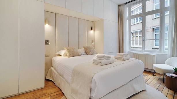 Une chambre moderne avec un grand lit, une armoire intégrée, une fenêtre donnant sur des immeubles et une petite table ronde agrémentée d’une plante.