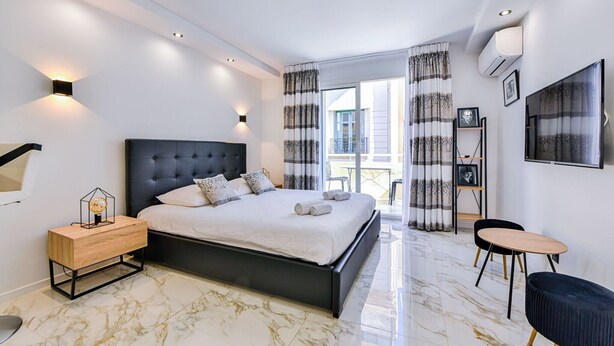 A modern bedroom with a large bed, bedside tables, a small table, and a chair. - Large studio apartment with terrace and sea view (Cannes)