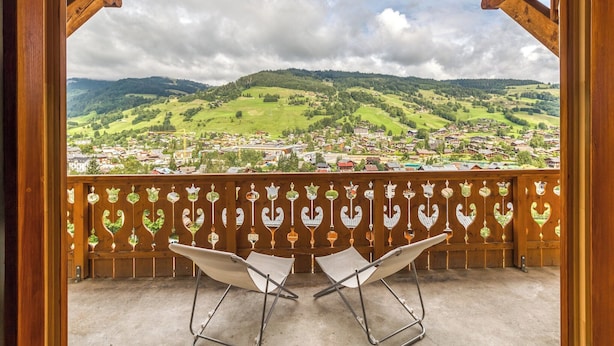 A balcony with two white chairs and a view of a green valley with houses.