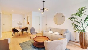 A modern living room with a sofa, coffee table, and chairs. There is a dining table with chairs and a potted plant. - Sleek apartment in Levallois-Perret (Levallois-Perret)