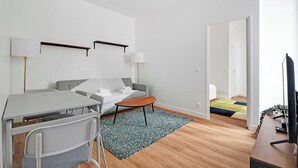 Ein modernes, minimalistisches Wohnzimmer mit einem grauen Sofa, einem kleinen Holztisch und einem grauen Sessel. Ein Fernseher steht auf einer Holzablage und eine Lampe auf einem Tisch.