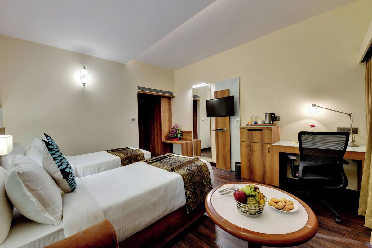 A hotel room with two beds, a desk, a chair, a TV, and a small dining table with fruit and cookies.