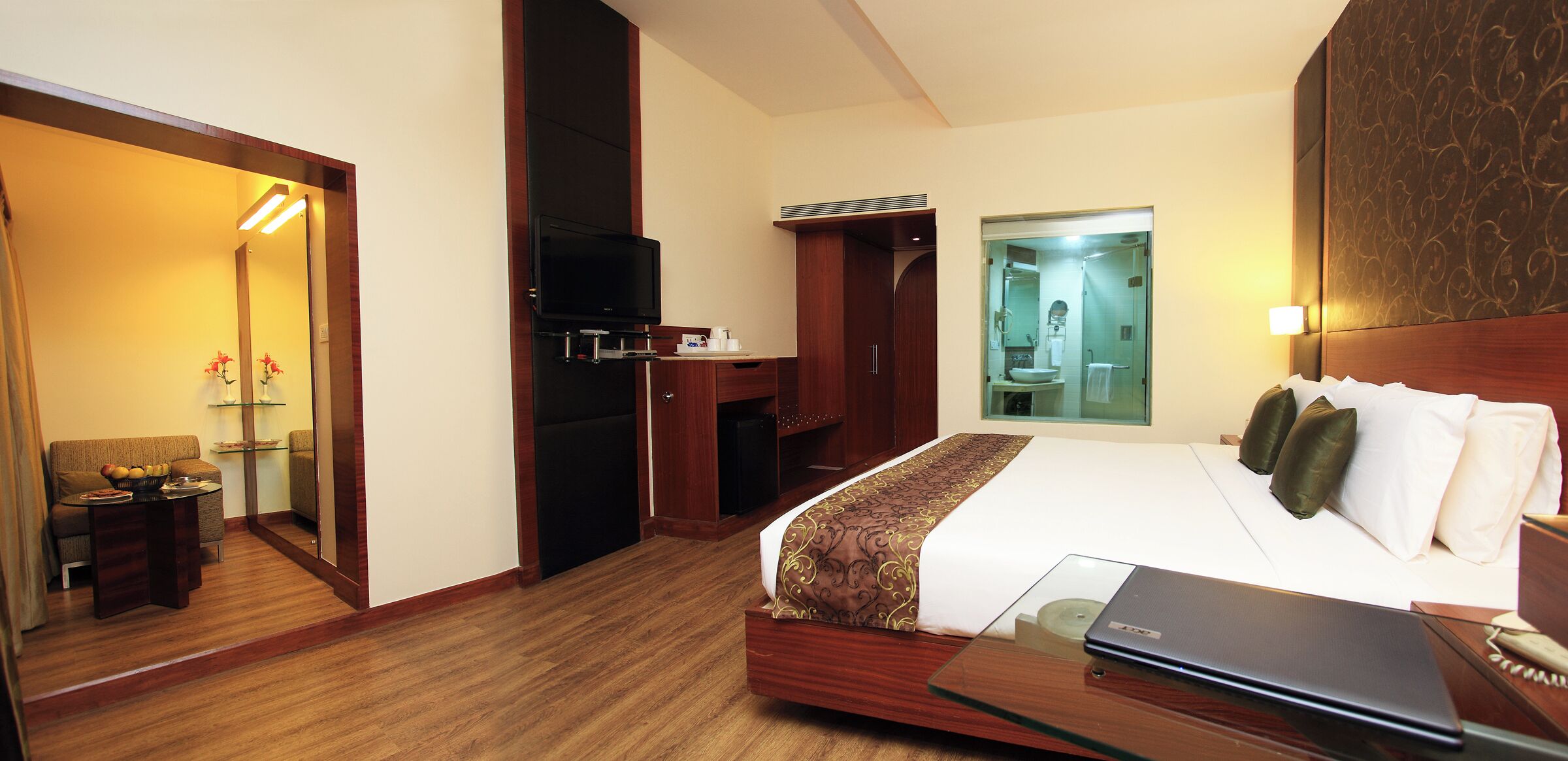 A hotel room with a bed, a desk, a television, and a bathroom visible through a glass door.