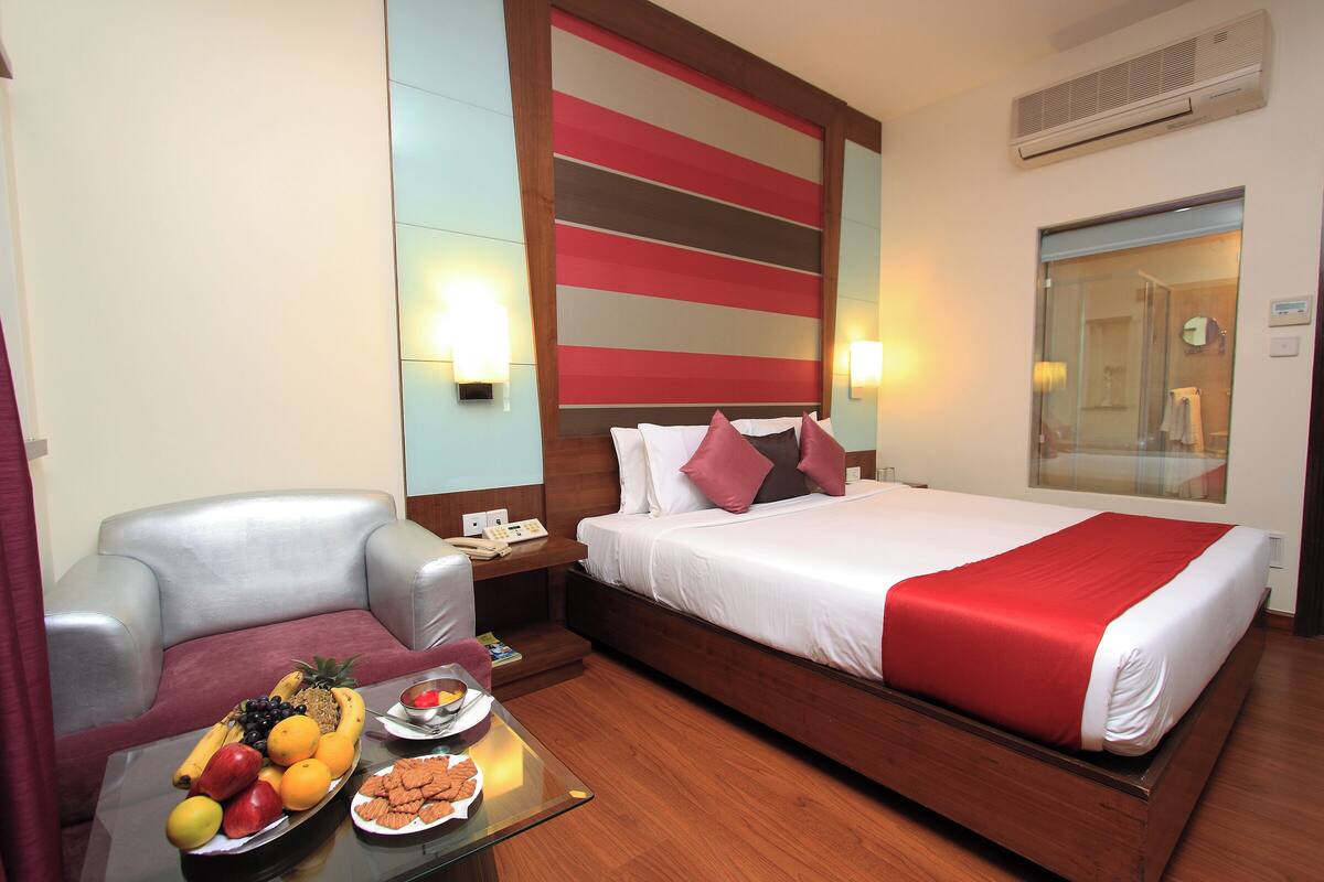 A hotel room with a bed, a sofa, a table with fruit and snacks, and a bathroom visible through a glass door.