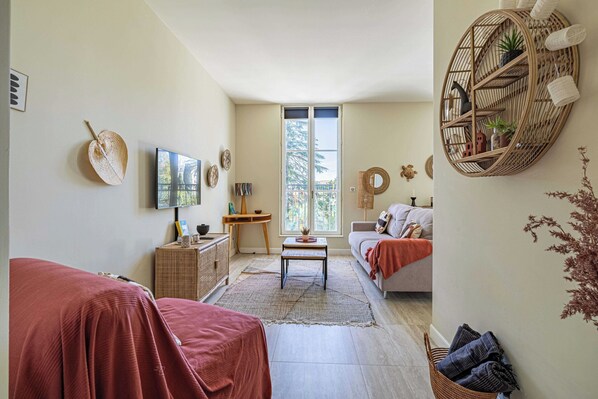 A cozy living room with a sofa, a coffee table, and a TV. There is a window with a view of trees and a balcony. - Quiet and luxurious studio - Cannes Center - 2p (Cannes)