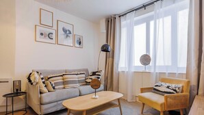 A modern living room with a sofa, armchair, coffee table, and a lamp. There are framed pictures on the wall and a window with curtains.