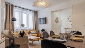 A modern living room with a sofa, coffee table, and dining table set for six. A television is mounted on the wall, and there are decorative wall pieces.