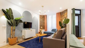 Una sala de estar moderna con un sofá, una mesa de centro y un televisor. Se observa una pared de ladrillo y una planta grande.
