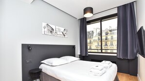 Ett modernt hotellrum med en stor säng, ett litet sidobord, en svart sänggavel, ett fönster med gardiner och utsikt över staden.