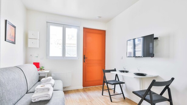 Ein modernes Wohnzimmer mit einem grauen Sofa, einem Esstisch mit zwei Stühlen, einem an der Wand befestigten Fernseher und einer leuchtend orangefarbenen Tür.