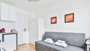 Ein modernes Wohnzimmer mit einer grauen Couch, einer kleinen Küchenzeile und zwei gerahmten Kunstwerken an der Wand.