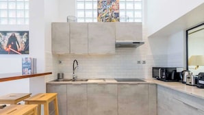 A modern kitchen with light-colored cabinets, a stainless steel sink, and a microwave. There are wooden stools and a window with artwork above the sink.