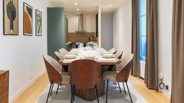 A modern dining room with a wooden table set for six, surrounded by six chairs. There are three framed artworks on the wall, a kitchen area with a modern stove, and a large window with curtains. - Magnificent apartment - 4BR/10P - Arc de Triomphe (Paris)