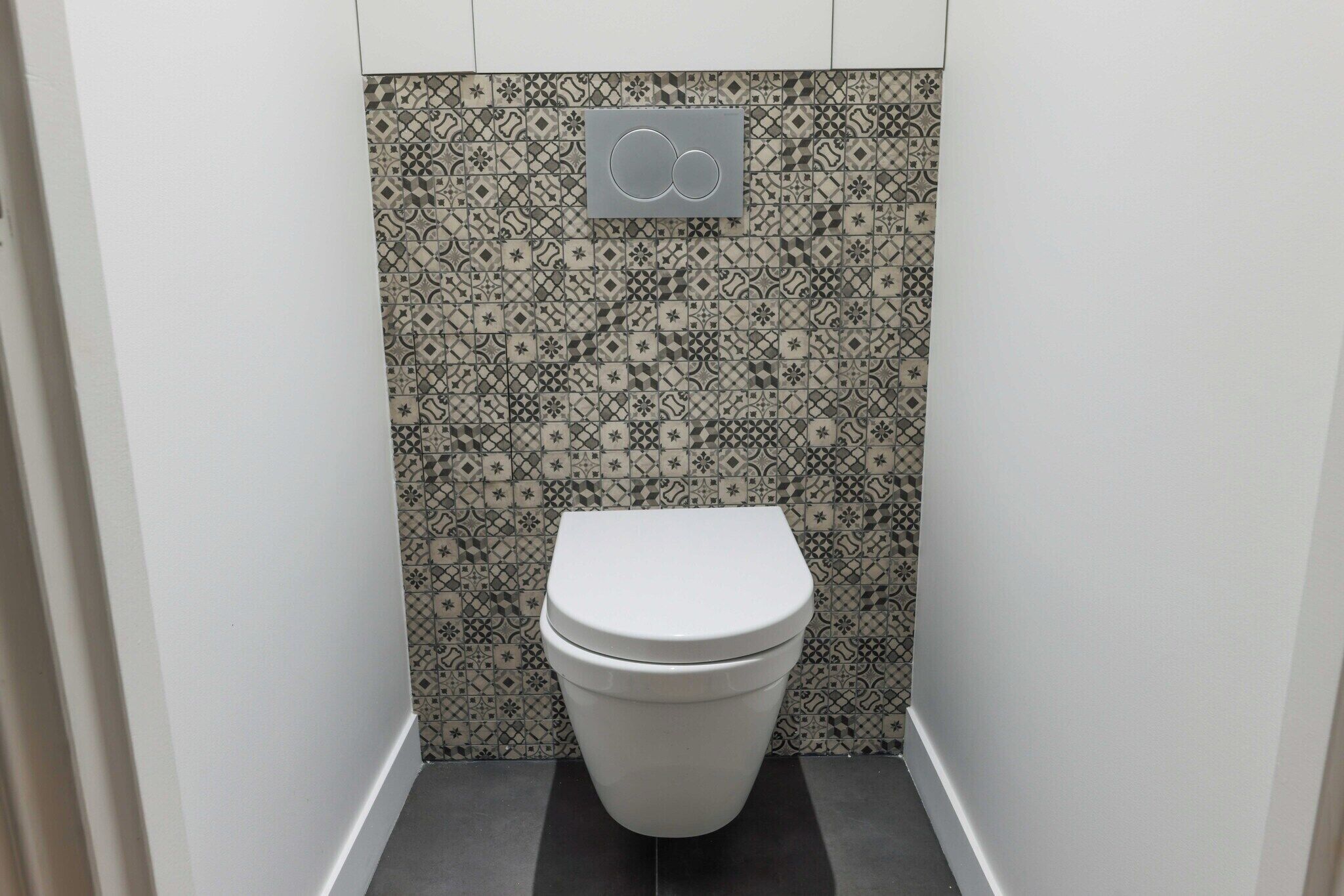 Une petite salle de bain moderne, dotée de toilettes, de carreaux décoratifs et d’une poignée de chasse d’eau fixée au mur.