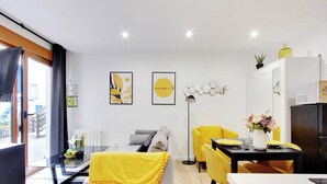 A modern living room with a sofa, a dining table with yellow chairs, and a kitchen area with a refrigerator and cabinets.