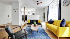 Uma sala de estar moderna com um sofá amarelo, poltronas pretas e um tapete azul. Há um ventilador de teto, uma área de jantar ao fundo e uma cozinha visível por uma porta aberta.