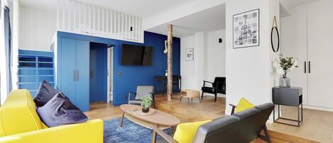 Una sala de estar moderna con un sofá amarillo, un sofá negro, una mesa de centro de madera y una alfombra azul. Hay un televisor montado en la pared, una mesita auxiliar con un florero y un cuadro enmarcado.