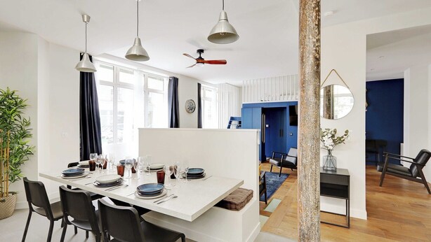 Uma sala de jantar moderna com uma mesa branca posta para seis pessoas, cadeiras pretas, luminárias pendentes e uma parede azul decorativa.
