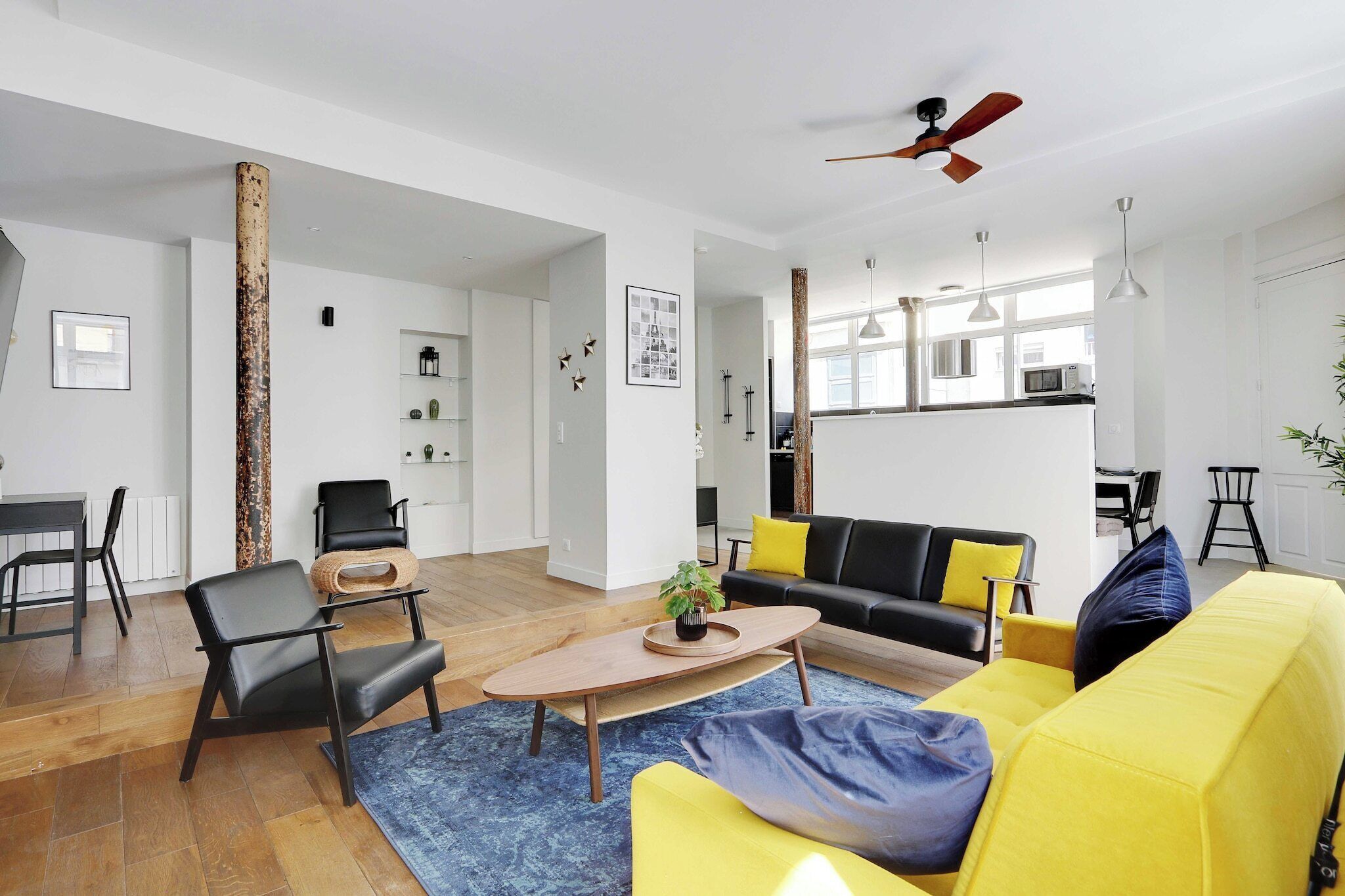 A spacious living area with a wooden coffee table, a yellow sofa, and a black sofa with yellow cushions. There is a black chair and a wooden pillar in the center.