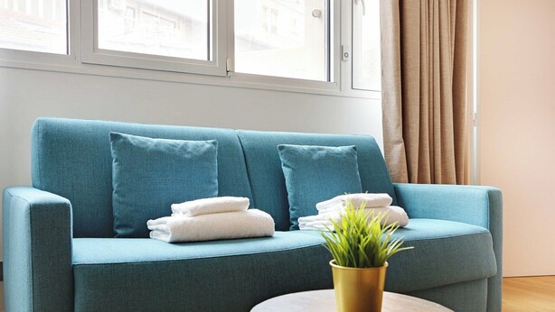 Ein blaugrünes Sofa mit weißen Handtüchern und einem Blumentopf, platziert in der Nähe eines Fensters mit Vorhängen.