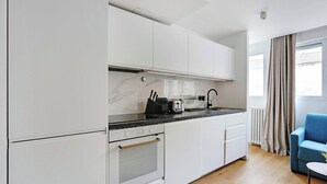 Eine moderne Küche mit weißen Schränken, einer schwarzen Arbeitsplatte und einem eingebauten Backofen. Ein Fenster mit Vorhängen und ein blauer Sessel sind im angrenzenden Wohnbereich zu sehen.
