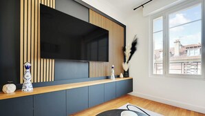 A modern living room with a large flat-screen TV mounted on a wall with vertical wooden slats. There is a wooden console table with decorative items, a large window with a view of buildings, and a black rug on the floor.