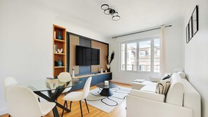 A modern living room with a glass dining table, white chairs, a sofa, and a TV mounted on a wooden panel wall.