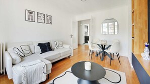A modern living room with a white sofa, a dining table with chairs, a coffee table, and a mirror. There are framed pictures on the wall and a ceiling light fixture.