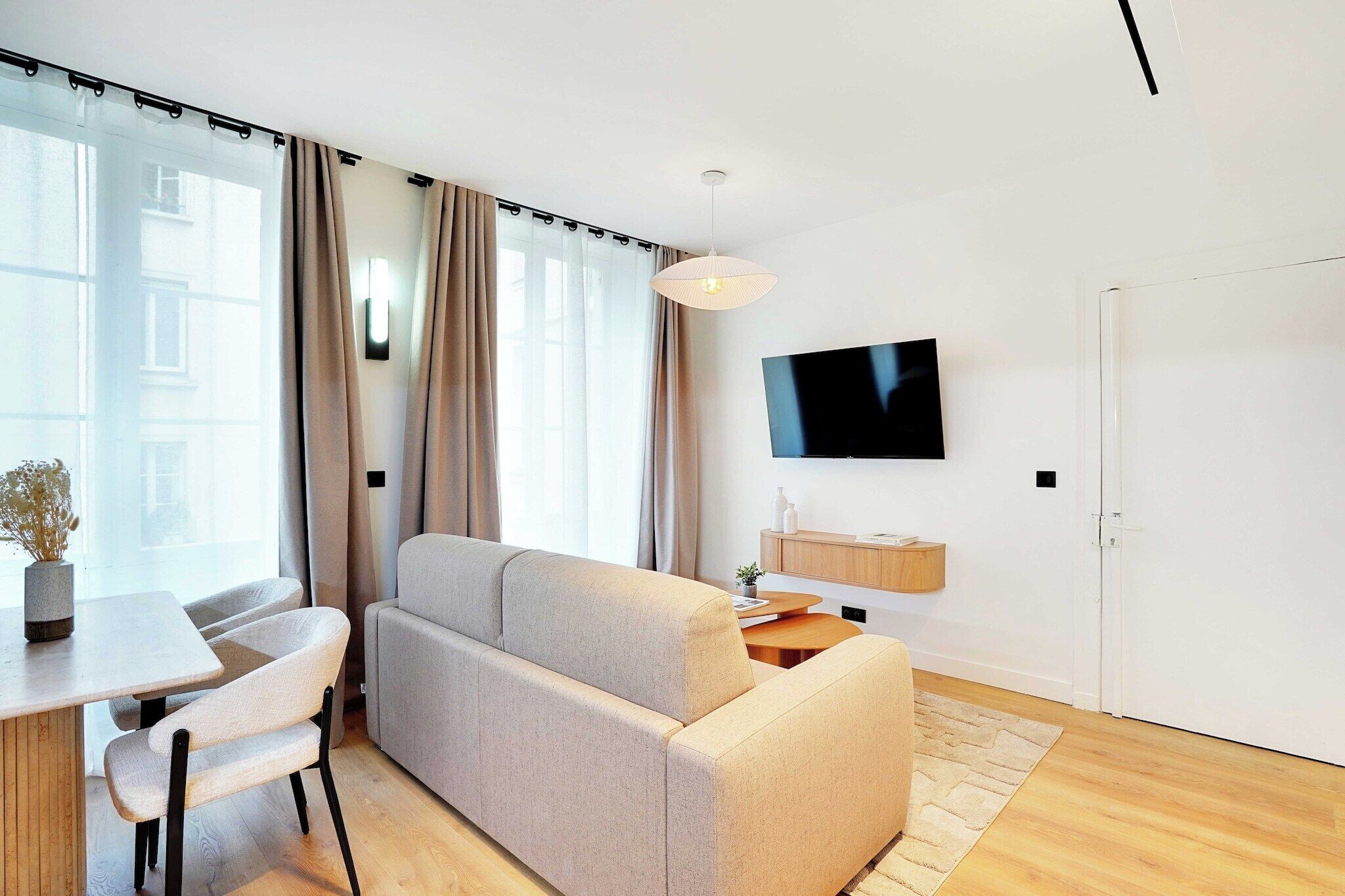 A modern living room with a sofa, a TV mounted on the wall, a dining table with chairs, and a large window with curtains.