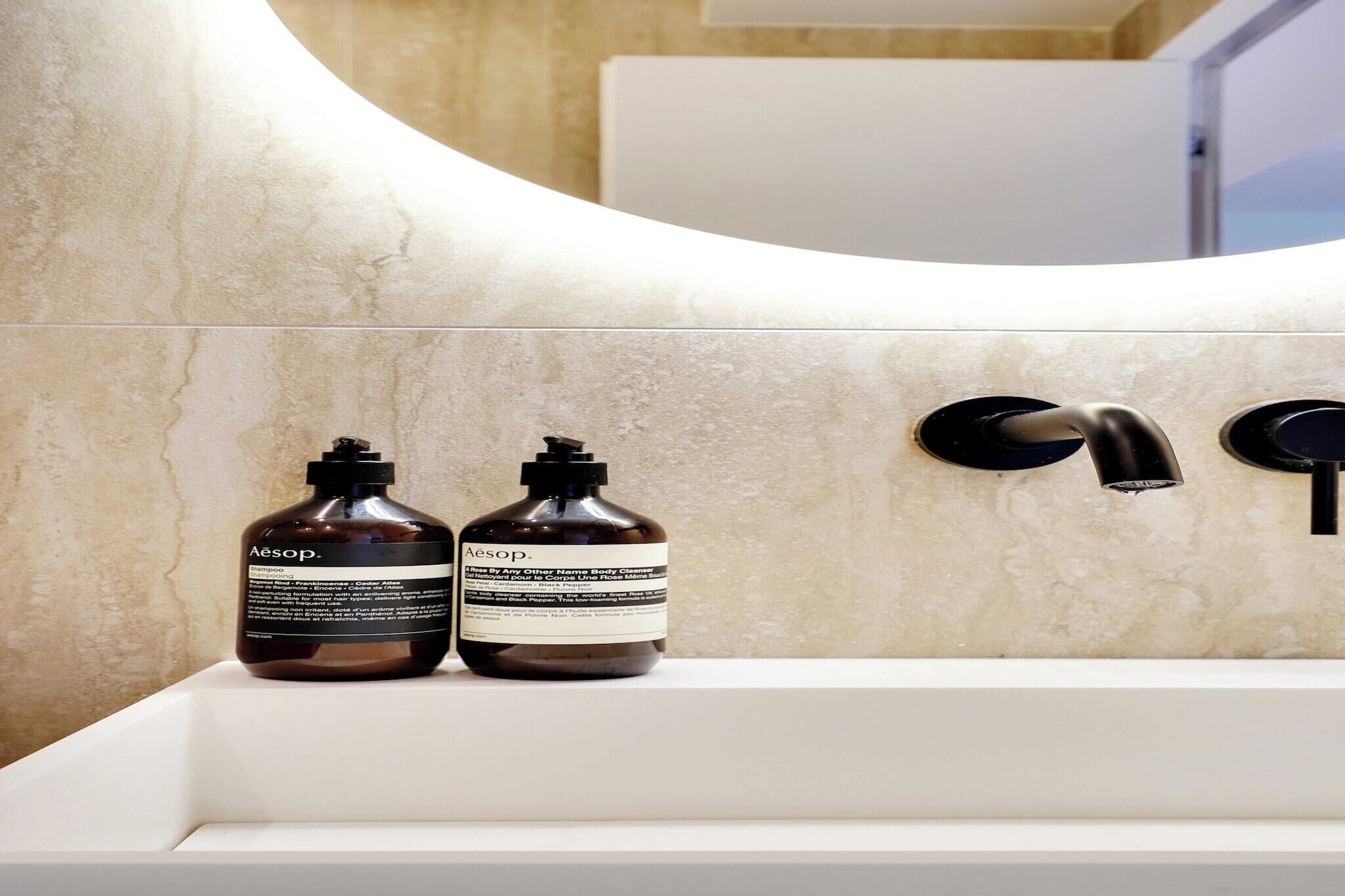 A bathroom with a white sink, two bottles of Aesop shampoo, and a black faucet.