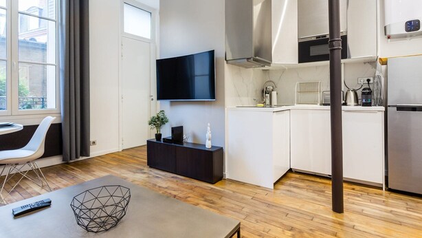 A modern living room with a flat-screen TV, a dark wooden coffee table, and a white sofa. The room has a large window with curtains, a kitchen area with white cabinets and stainless steel appliances, and a wooden floor.