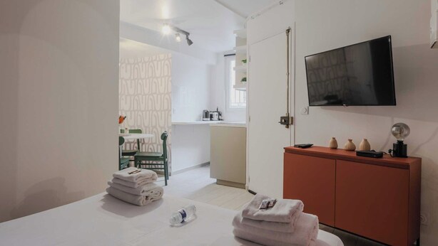 Una habitación de hotel con cama, televisor montado en la pared, un mueble con adornos y vista al baño por una puerta abierta.