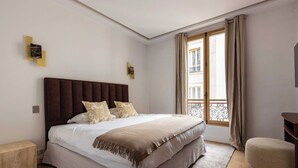 Un dormitorio ordenado con una cama grande, una mesita de noche, una silla y una ventana con cortinas.