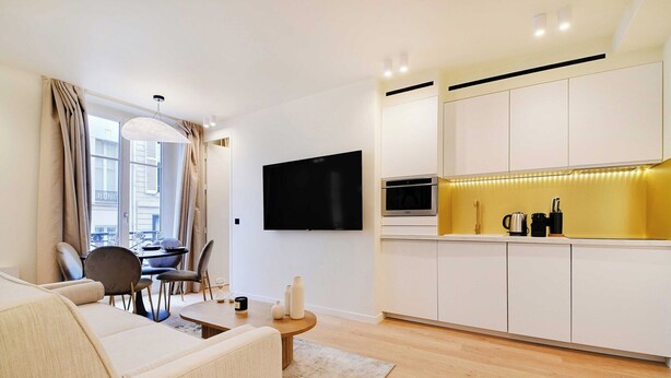A modern living room with a sofa, dining table, and chairs. A flat-screen TV is mounted on the wall. The kitchen features white cabinets and a yellow countertop.