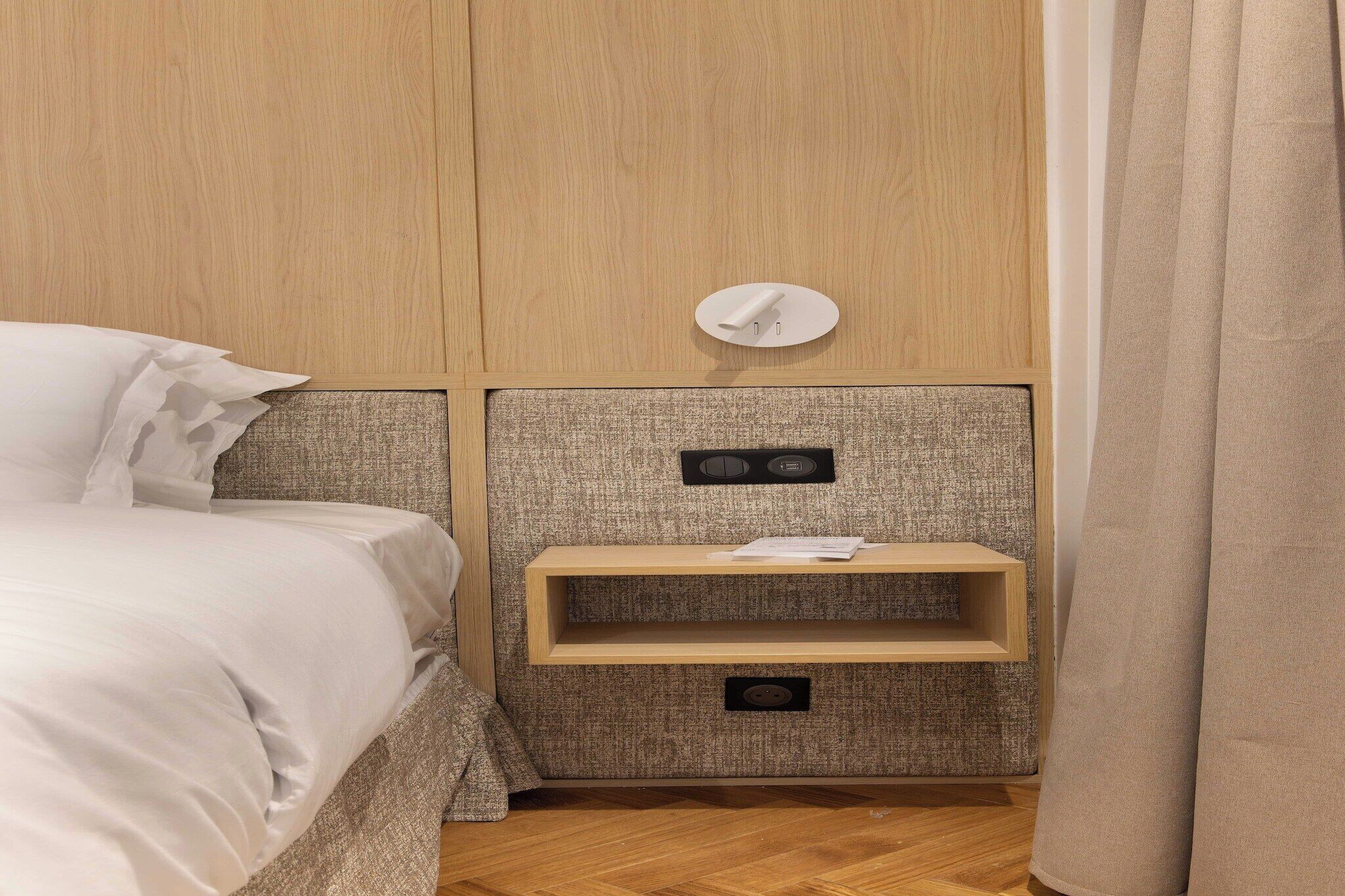 A neatly made bed with white linens, a wooden nightstand with a shelf, and a power outlet.