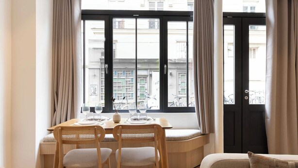 A dining area with a table set for two, a bench, and a view of the street through large windows. - Beautiful Apartment - 1BD/4P - Quartier Latin (Paris)