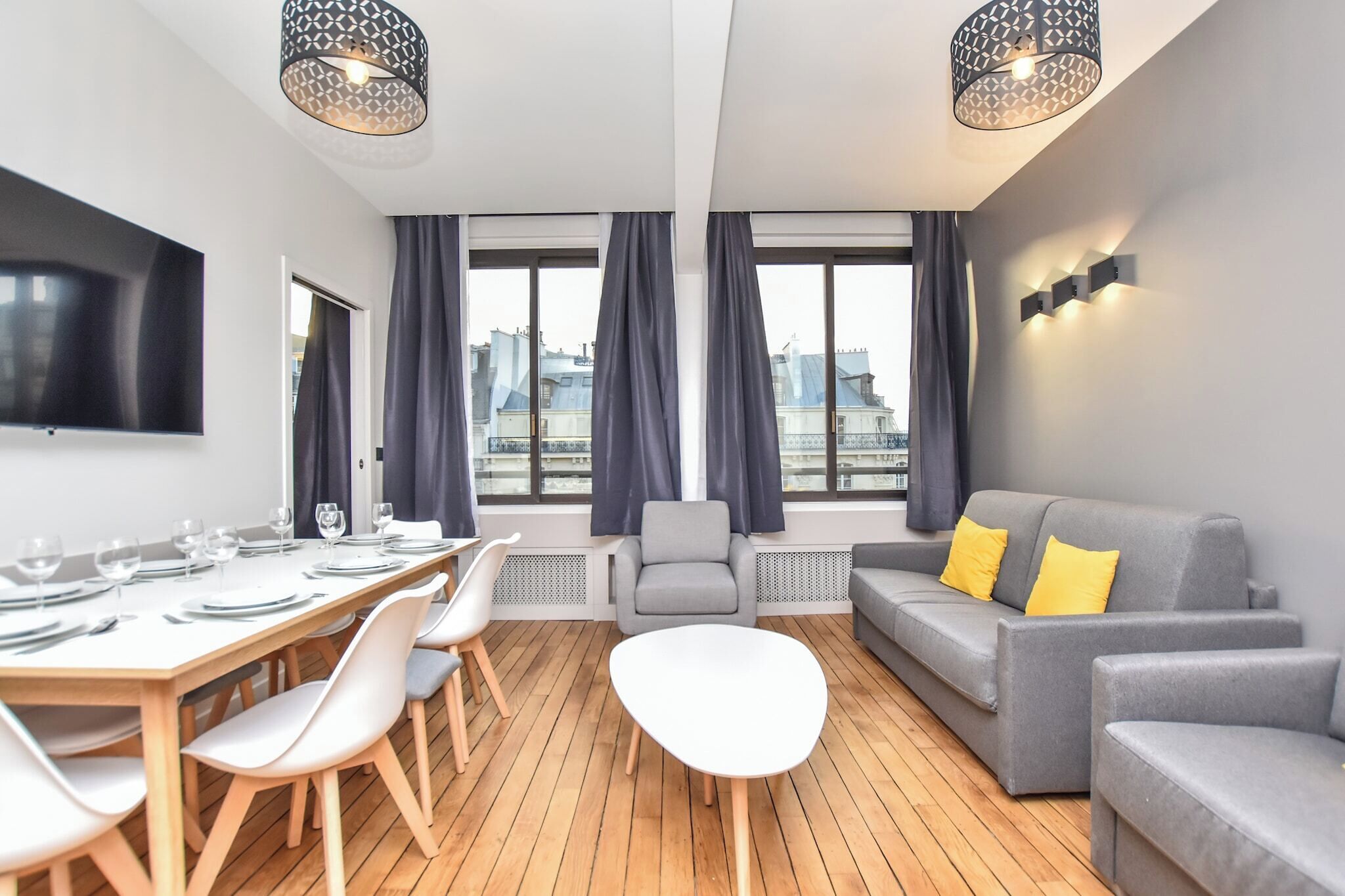 Un salon moderne comprenant un coin repas, des canapés gris, une table blanche et de grandes fenêtres avec des rideaux.