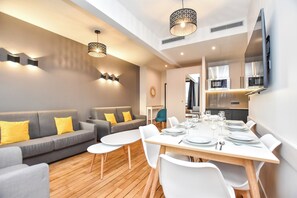 Un salon moderne avec un canapé gris, des coussins jaunes, une table à manger en bois dressée pour six personnes et des luminaires suspendus.