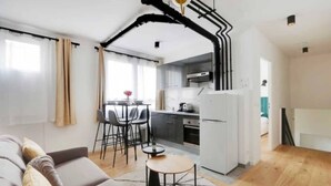 Een moderne woonkamer met een bank, een eethoek met stoelen, een keuken met een koelkast, en een trap met zwarte buizen.
