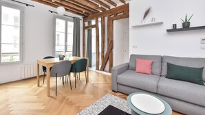 Un soggiorno moderno con un divano grigio, travi di legno al soffitto, un'area pranzo con tavolo e sedie, e ampie finestre.
